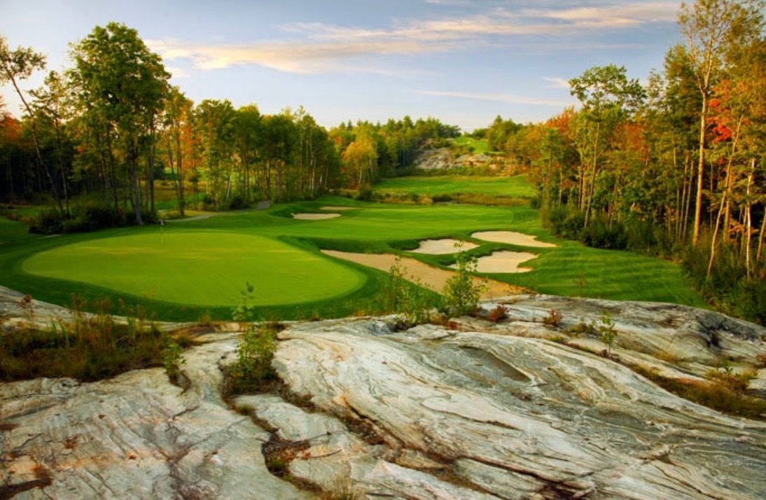 Scenic view of a lush golf course surrounded by trees and rocky terrain under a blue sky.