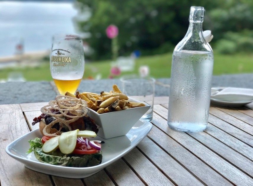 Burger with onion rings and fries, beer glass, and water bottle on a wooden table outdoors.